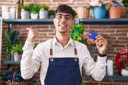 Photo for Young hispanic man working at florist shop holding credit card pointing thumb up to the side smiling happy with open mouth - Royalty Free Image