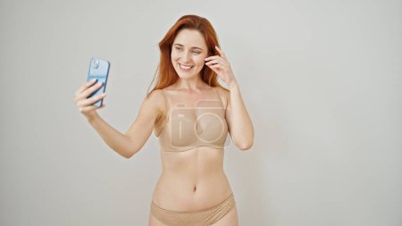 Foto de Mujer pelirroja joven con lencería hacer selfie por teléfono inteligente sonriendo sobre fondo blanco aislado - Imagen libre de derechos