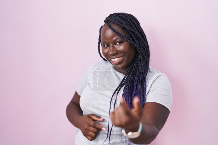Foto de Joven mujer africana de pie sobre fondo rosa señas vienen aquí gesto con la mano invitando a la bienvenida feliz y sonriente - Imagen libre de derechos