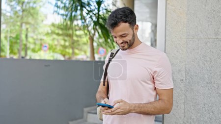 Téléchargez les photos : Jeune homme hispanique utilisant un smartphone portant un sac à dos dans la rue - en image libre de droit
