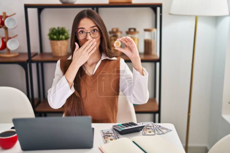 Téléchargez les photos : Jeune femme brune travaillant avec un ordinateur portable tenant monnaie virtuelle Bitcoin couvrant la bouche avec la main, choqué et peur de l'erreur. expression surprise - en image libre de droit