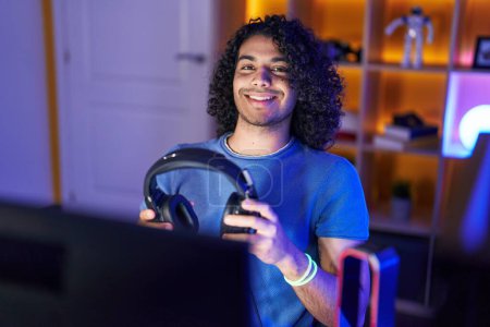 Photo for Young latin man streamer smiling confident holding headphones at gaming room - Royalty Free Image