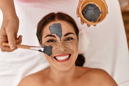 Photo for Young beautiful hispanic woman lying on table having facial treatment at beauty salon - Royalty Free Image