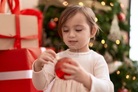Photo for Adorable girl holding ball decoration standing by christmas tree at home - Royalty Free Image
