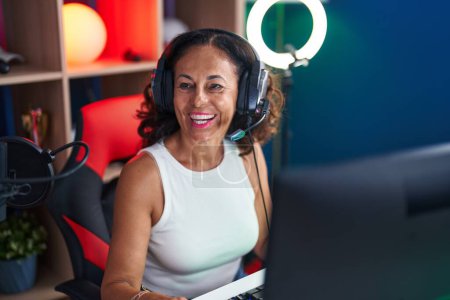Téléchargez les photos : Middle age woman streamer smiling confident sitting on table at gaming room - en image libre de droit