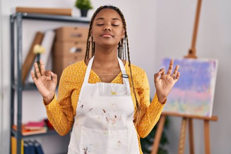 Téléchargez les photos : Femme afro-américaine avec des tresses au studio d'art se détendre et sourire les yeux fermés en faisant geste de méditation avec les doigts. concept de yoga. - en image libre de droit