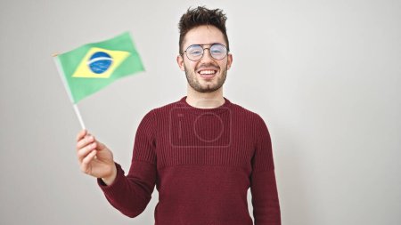 Foto de Joven hombre hispano sonriendo confiado sosteniendo bandera brasileña sobre fondo blanco aislado - Imagen libre de derechos
