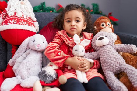 Photo for Adorable hispanic girl hugging teddy bear sitting on sofa by christmas decoration at home - Royalty Free Image