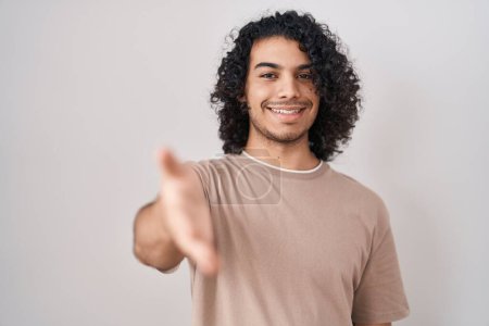 Foto de Hombre hispano con el pelo rizado de pie sobre fondo blanco sonriendo amistoso ofreciendo apretón de manos como saludo y bienvenida. negocios exitosos. - Imagen libre de derechos