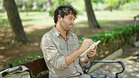 Téléchargez les photos : Jeune homme hispanique écrivant sur un cahier assis sur un banc pensant au parc - en image libre de droit