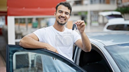 Téléchargez les photos : Jeune homme hispanique tenant la clé de la nouvelle voiture penchée sur la voiture de porte à la rue - en image libre de droit