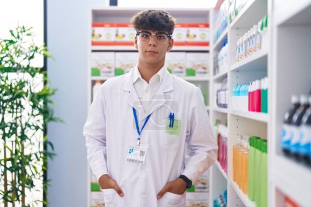 Téléchargez les photos : Jeune adolescent hispanique pharmacien debout avec une expression détendue à la pharmacie - en image libre de droit