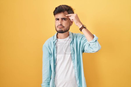Photo for Young hispanic man with tattoos standing over yellow background pointing unhappy to pimple on forehead, ugly infection of blackhead. acne and skin problem - Royalty Free Image