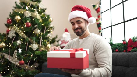 Photo for Young hispanic man holding gift sitting on sofa by christmas tree at home - Royalty Free Image