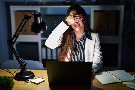Téléchargez les photos : Jeune femme brune travaillant au bureau la nuit avec un ordinateur portable couvrant les yeux avec la main, l'air grave et triste. concept de non-visibilité, de dissimulation et de rejet - en image libre de droit
