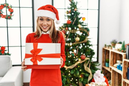 Photo for Young caucasian woman unpacking gift standing by christmas tree at home - Royalty Free Image