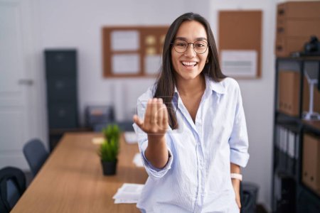Téléchargez les photos : Jeune femme hispanique au bureau faisant signe de venir ici geste avec la main invitant accueillant heureux et souriant - en image libre de droit