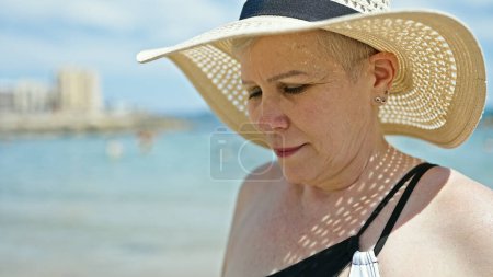 Photo for Middle age grey-haired woman tourist wearing swimsuit and summer hat looking down at the beach - Royalty Free Image