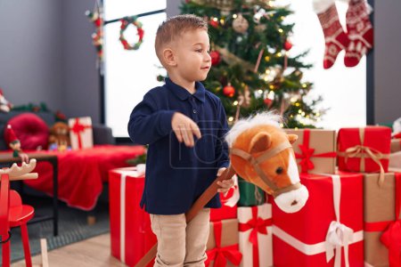 Photo for Adorable hispanic boy playing with horse toy standing by christmas tree at home - Royalty Free Image