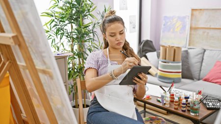 Photo for Stunning young hispanic female artist caught in a moment of inspiration, gleefully drawing on touchpad amidst the canvas, brushes and paint palette in her art studio - Royalty Free Image