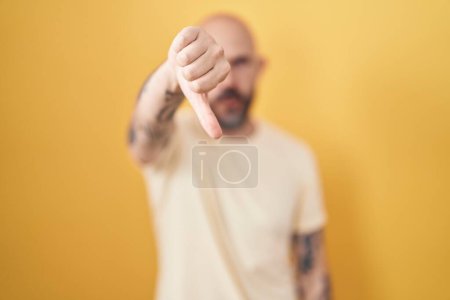 Téléchargez les photos : Homme hispanique avec des tatouages debout sur fond jaune regardant malheureux et en colère montrant rejet et négatif avec les pouces vers le bas geste. mauvaise expression. - en image libre de droit