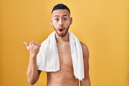 Téléchargez les photos : Jeune homme hispanique debout torse nu avec serviette surprise pointant avec le doigt de la main sur le côté, bouche ouverte expression étonnante. - en image libre de droit