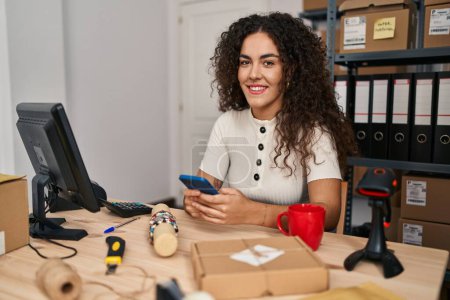 Foto de Joven mujer hispana hermosa comercio electrónico trabajador de negocios usando teléfono inteligente beber café en la oficina - Imagen libre de derechos