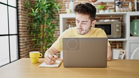 Photo for Young hispanic man student using laptop taking notes at dinning room - Royalty Free Image