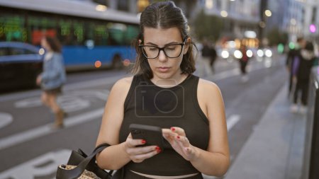Téléchargez les photos : Belle femme hispanique brune à lunettes, absorbée par une conversation sérieuse sur smartphone dans les rues urbaines de Madrid - en image libre de droit