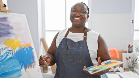 Téléchargez les photos : Joyeux artiste afro-américaine, tenant avec confiance le pinceau et la palette, souriant lumineux dans le studio d'art d'intérieur - en image libre de droit