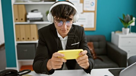 Téléchargez les photos : Chillaxed jeune hispanique travail patron adolescent jouer à des jeux vidéo à l'intérieur au bureau, à l'écoute avec des écouteurs, absorbé dans le monde du joueur, le bruit de fond de l'agitation des affaires s'estompe! - en image libre de droit