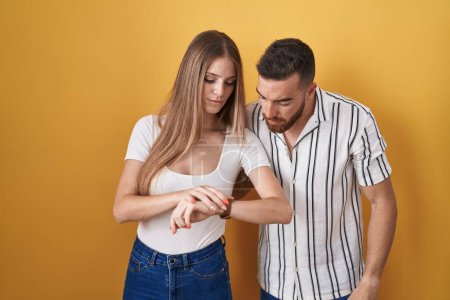 Photo for Young couple standing over yellow background checking the time on wrist watch, relaxed and confident - Royalty Free Image