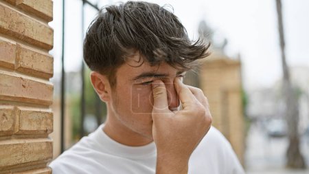 Photo for Handsome young hispanic teenager rubbing itchy eyes on sunlit city street, serious expression portraying urban lifestyle stress - Royalty Free Image