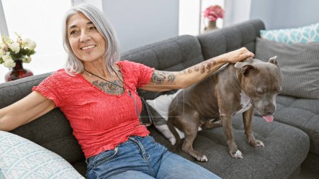 Téléchargez les photos : Joyeuse femme d'âge moyen assise sur le canapé à la maison, jouant avec son chien bien-aimé aux cheveux gris, riant et exprimant un pur bonheur - en image libre de droit