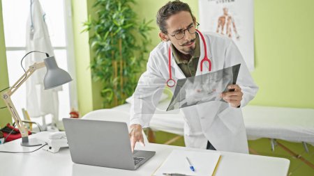 Téléchargez les photos : Jeune homme hispanique médecin regardant les rayons X à l'aide d'un ordinateur portable à la clinique - en image libre de droit