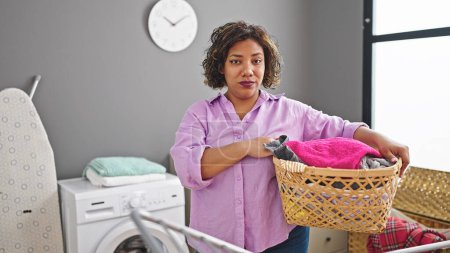 Photo for Young beautiful latin woman holding basket with clothes with serious face at laundry room - Royalty Free Image