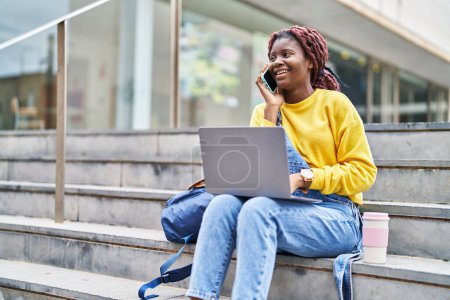 Téléchargez les photos : Étudiante afro-américaine utilisant un ordinateur portable parlant sur smartphone à l'université - en image libre de droit