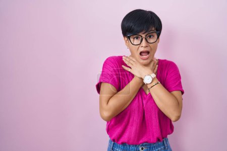 Foto de Mujer asiática joven con el pelo corto de pie sobre fondo rosa gritando sofocar porque estrangulamiento doloroso. problema de salud. concepto de asfixia y suicidio. - Imagen libre de derechos