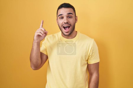 Téléchargez les photos : Jeune homme hispanique debout sur fond jaune pointant le doigt vers le haut avec une idée réussie. Sorti et heureux. numéro un. - en image libre de droit