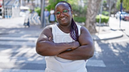 Foto de Mujer afroamericana confiada, trenzas deportivas y gafas, sonriendo ampliamente y de pie con los brazos cruzados en una calle urbana, irradiando alegría positiva y expresión al aire libre. - Imagen libre de derechos