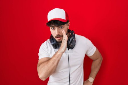 Foto de Hombre hispano con barba usando sombrero de gamer y auriculares mano sobre boca contando rumores secretos, susurrando conversación maliciosa - Imagen libre de derechos