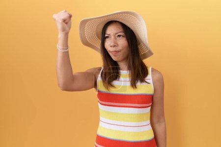 Foto de Mujer china de mediana edad con sombrero de verano sobre fondo amarillo enojado y loco levantando el puño frustrado y furioso mientras gritaba con ira. rabia y concepto agresivo. - Imagen libre de derechos