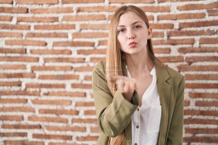 Foto de Young caucasian woman standing over bricks wall background looking at the camera blowing a kiss with hand on air being lovely and sexy. love expression. - Imagen libre de derechos