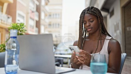 Foto de Mujer afroamericana usando portátil y teléfono inteligente sentado en la mesa en la terraza de la cafetería - Imagen libre de derechos
