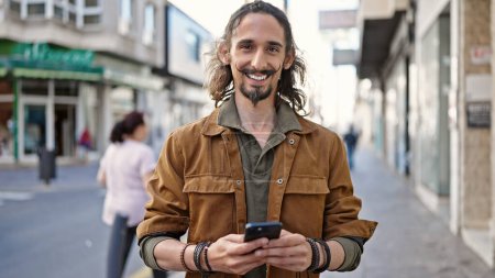 Photo for Young hispanic man using smartphone smiling at street - Royalty Free Image