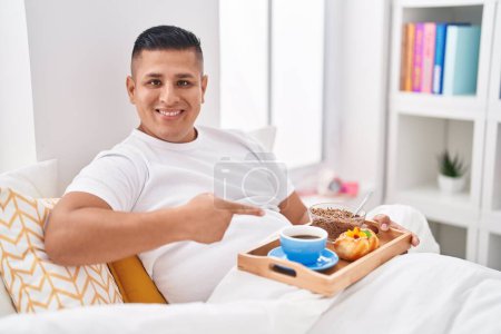 Foto de Joven hispano desayunando en la cama sonriendo feliz señalando con la mano y el dedo - Imagen libre de derechos