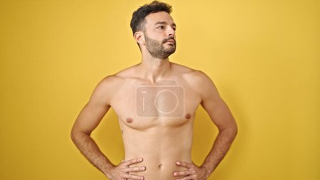Téléchargez les photos : Jeune homme hispanique touriste debout torse nu regardant sur le côté avec un visage sérieux sur fond jaune isolé - en image libre de droit