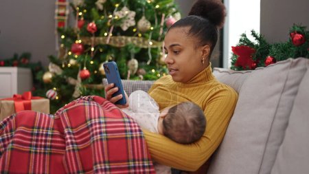 Photo for Mother and son celebrating christmas using smartphone while breastfeeding baby at home - Royalty Free Image