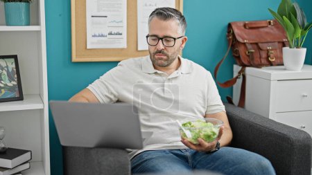 Photo for Grey-haired man business worker using laptop eating salad at the office - Royalty Free Image
