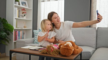 Photo for Smiling caucasian mother and little daughter, sitting on sofa at home, making memories together, drawing on notebook and taking fun-filled selfie picture with phone. - Royalty Free Image